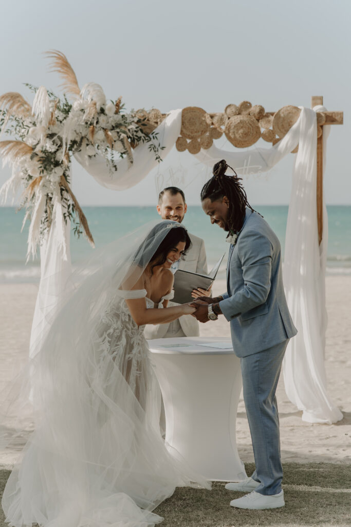 Bilingual wedding officiant for Puerto Rico beach weddings and elopements. A bride and groom share a joyful moment during a beach wedding. The bride laughs while the groom smiles, under a floral arch with the ocean behind. 