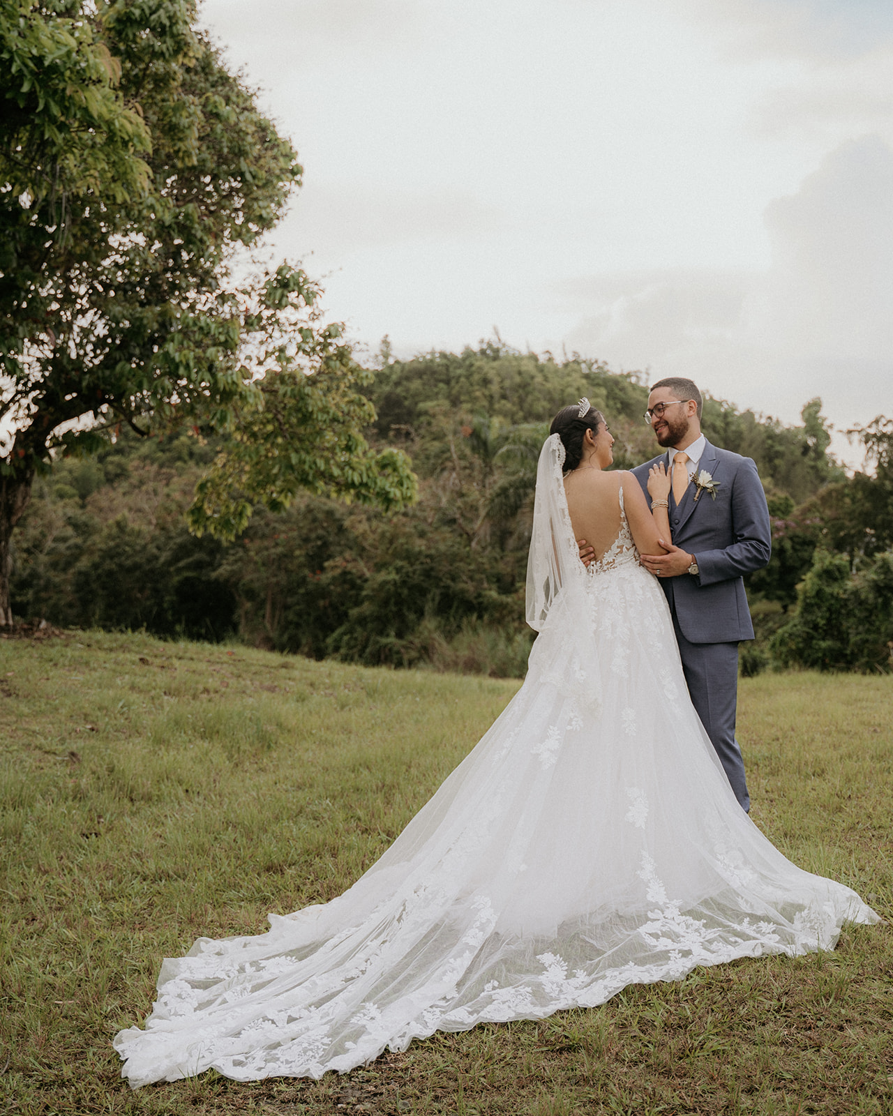 Bride in a flowing white gown and groom in blue suit stand smiling on lush grass, near a tree, exuding joy against a serene, cloudy sky backdrop.