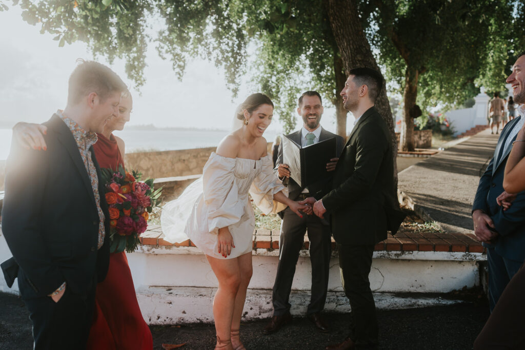 A joyful couple holds hands during an outdoor wedding ceremony in Old San Juan, Puerto Rico. Zevio officiates, while guests, dressed in formal attire, smile nearby under lush trees.