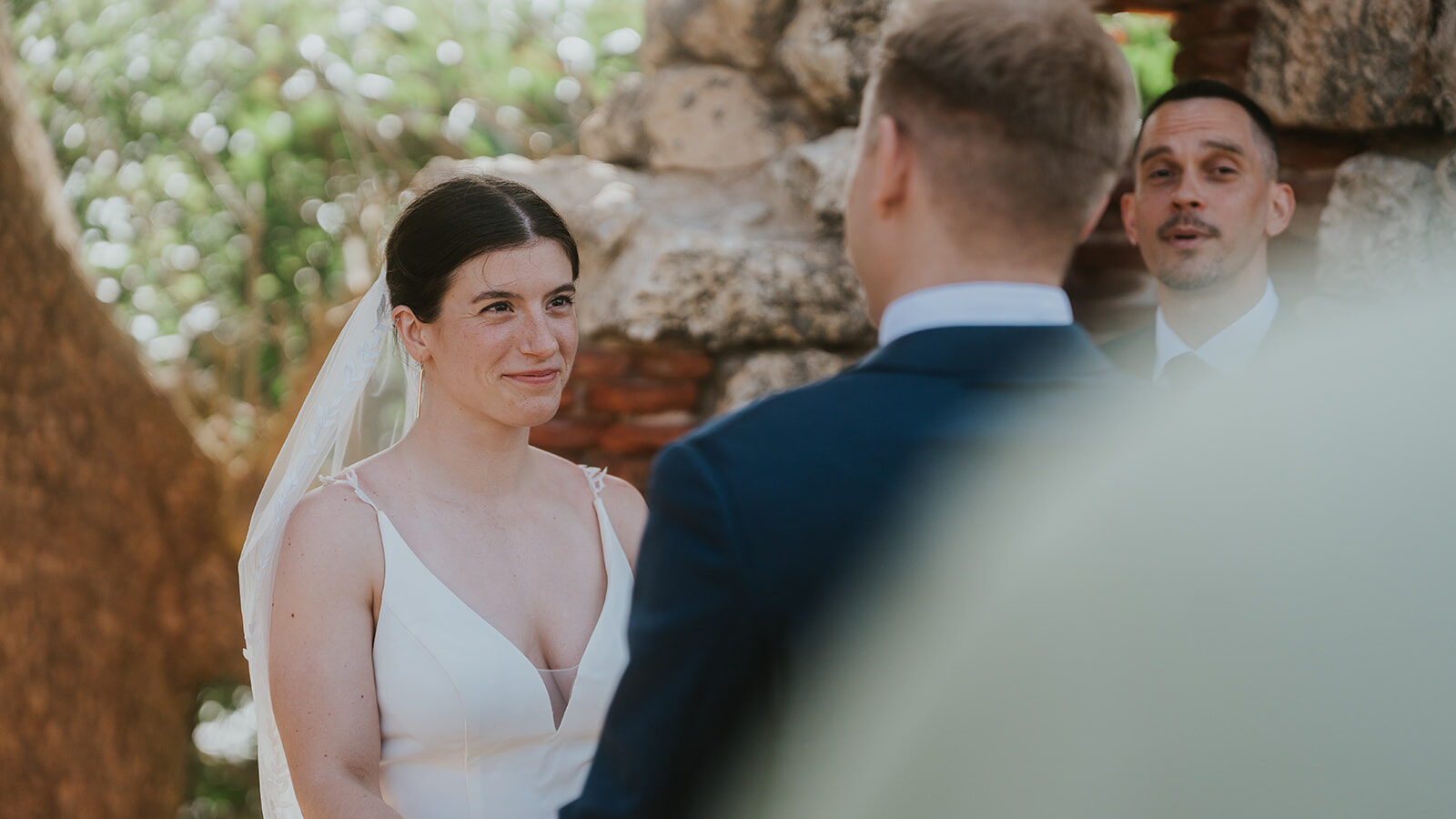 Zevio officiating an outdoor elopement ceremony in Aguadilla, Puerto Rico. Bride and groom hold hands, facing each other. The bride wears a white dress and veil, smiling gently. The groom stands in a dark suit.