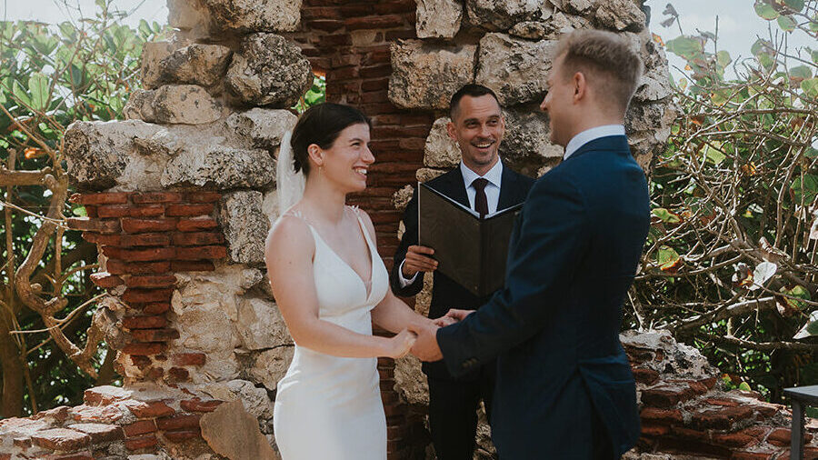 Zevio officiates an outdoor elopement ceremony in Aguadilla, Puerto Rico.  A bride in a white gown and groom in a navy suit hold hands, smiling.  Stone ruins and foliage surround them under a bright sky.